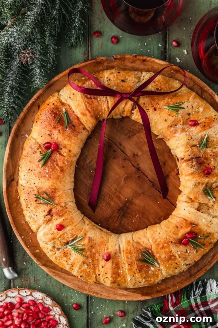 Flaky Bacon and Brie Crescent Wreath Recipe 12 A wood board with a bacon brie crescent wreath on a green counter next to garland, a plaid linen, glasses of red wine, and a white plate of pomegranate arils.