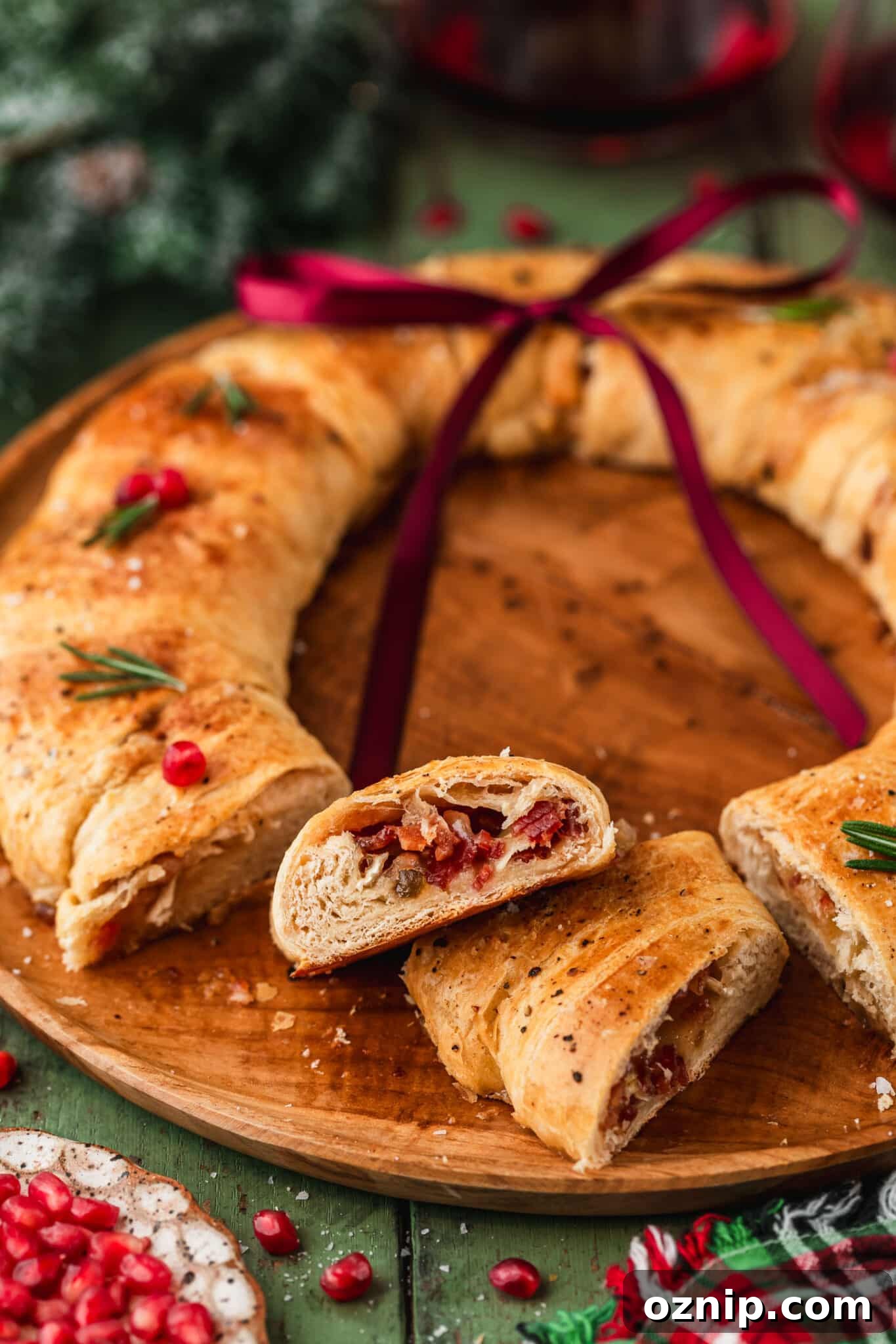 Flaky Bacon and Brie Crescent Wreath Recipe 13 A sliced bacon brie crescent wreath on a wood board next to glasses of red wine, garland, and a plaid linen on a green table.