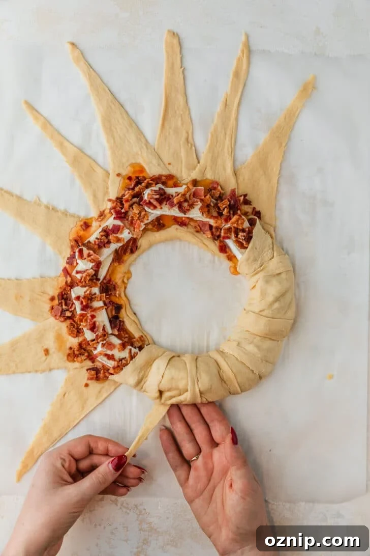 Flaky Bacon and Brie Crescent Wreath Recipe 8 Hands wrapping crescent dough around bacon and cheese on parchment paper.