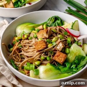 Sesame-Ginger Tofu Soba Noodles 11 A white bowl filled with light-brown soba noodles, golden crispy tofu cubes, bright green edamame, and glossy sautéed bok choy, sprinkled with sliced green onions.