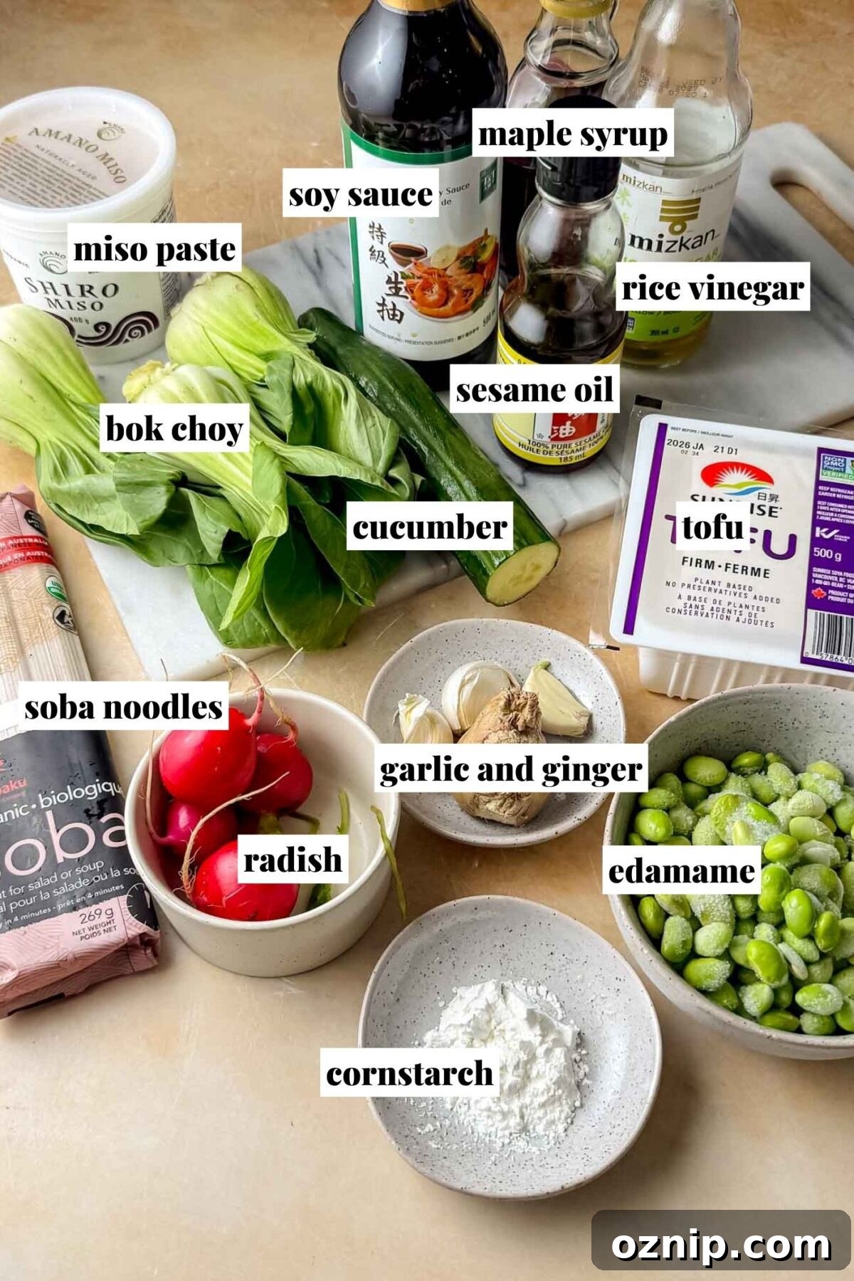 Sesame-Ginger Tofu Soba Noodles 3 A countertop display of ingredients for tofu soba noodles, including bottles of soy sauce, maple syrup, rice vinegar, and sesame oil, a tub of white miso paste, fresh bok choy, a whole cucumber, a package of firm tofu, frozen edamame, dry soba noodles, red radishes, fresh garlic and ginger, and a small bowl of cornstarch arranged on a cutting board and in bowls.