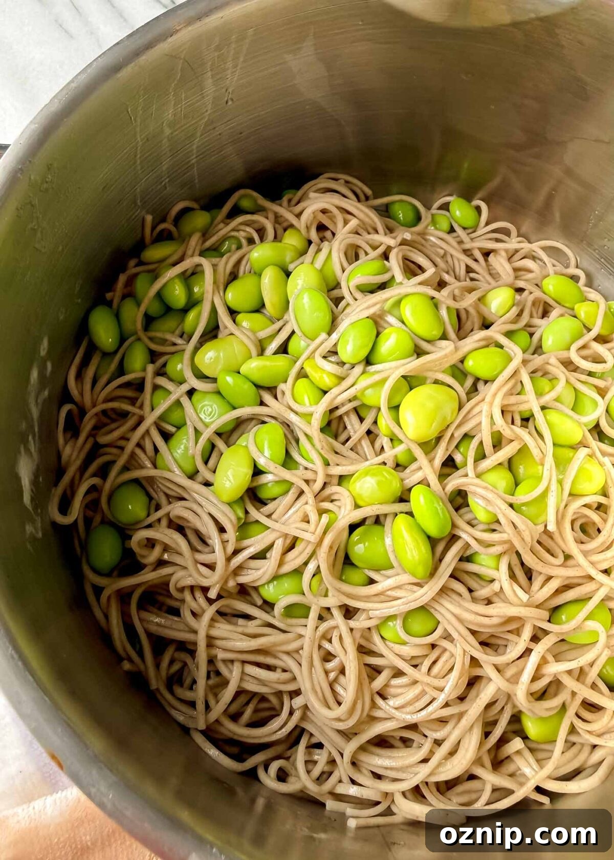 Sesame-Ginger Tofu Soba Noodles 7 A stainless steel pot filled with cooked soba noodles tangled together with bright green edamame beans, viewed from above so the glossy noodles and beans fill the frame.