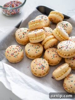 A batch of birthday cake macarons on crumpled parchment paper, with rainbow nonpareil sprinkles on and beside them.
