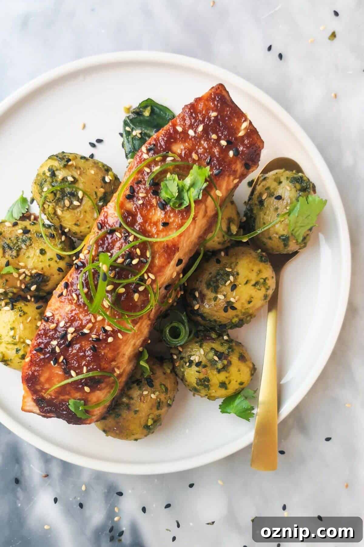 Miso glazed salmon on top of herby potatoes on a white plate, with sesame seeds, coriander and spring onion on top.