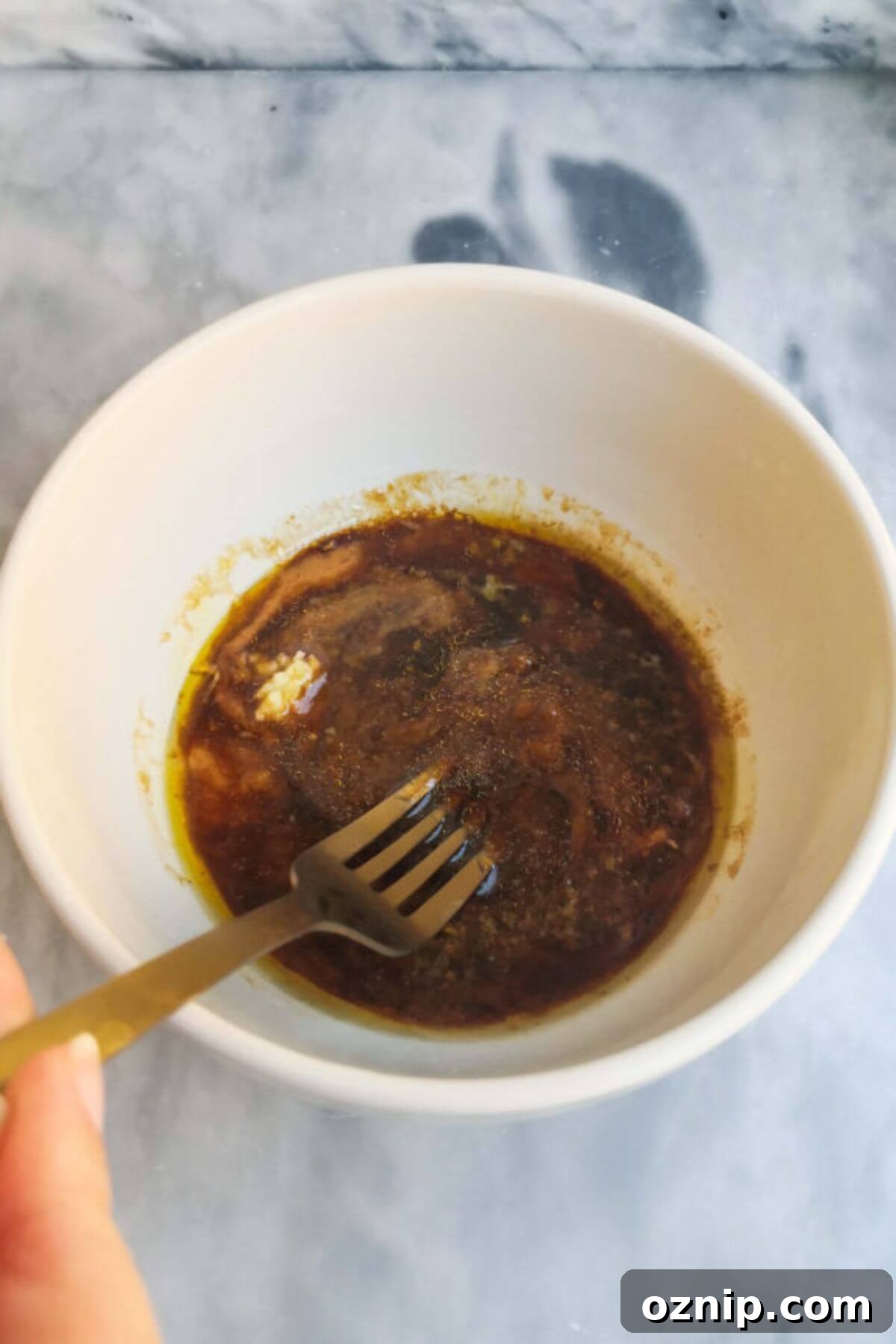 A gold fork mixing miso marinade in a small white bowl on a grey marble background.