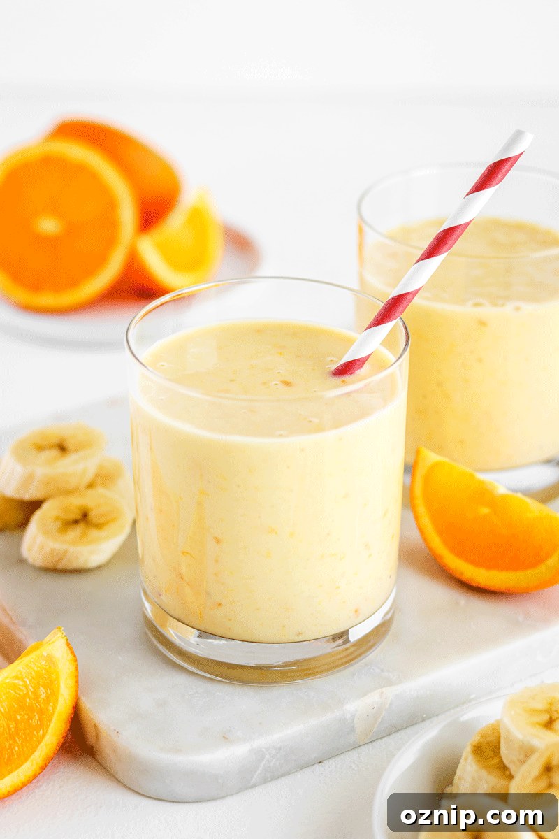 A glass of orange smoothie with a red striped straw with orange slices and banana slices around it.