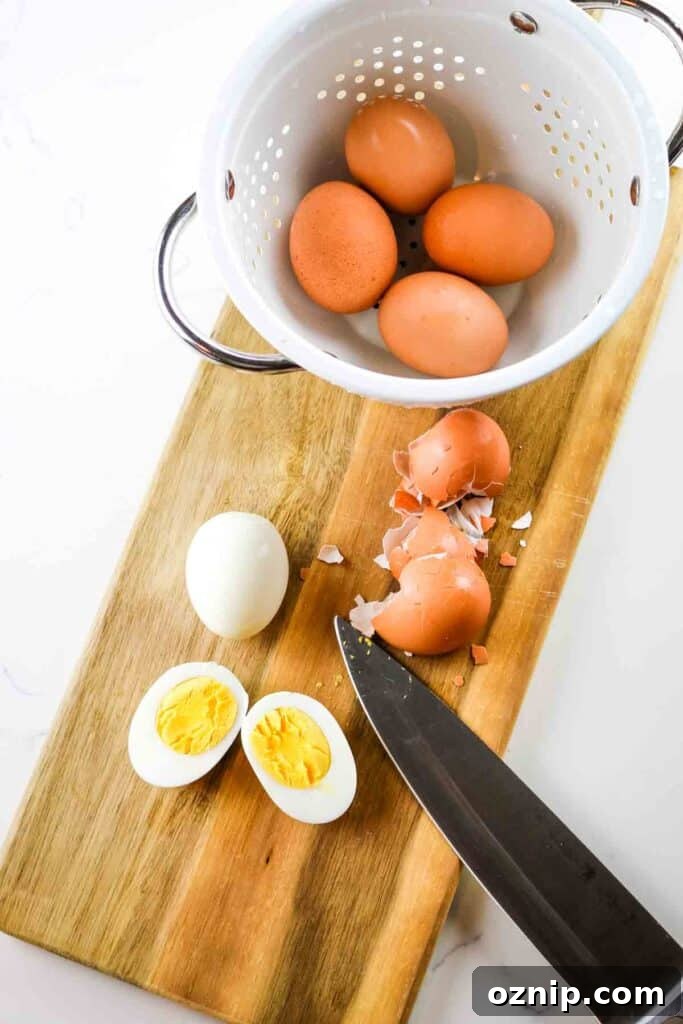 Classic Deviled Eggs Recipe for Parties 3 Hard boiled eggs on a cutting board with knife and colander, with one egg peeled and cut in half.