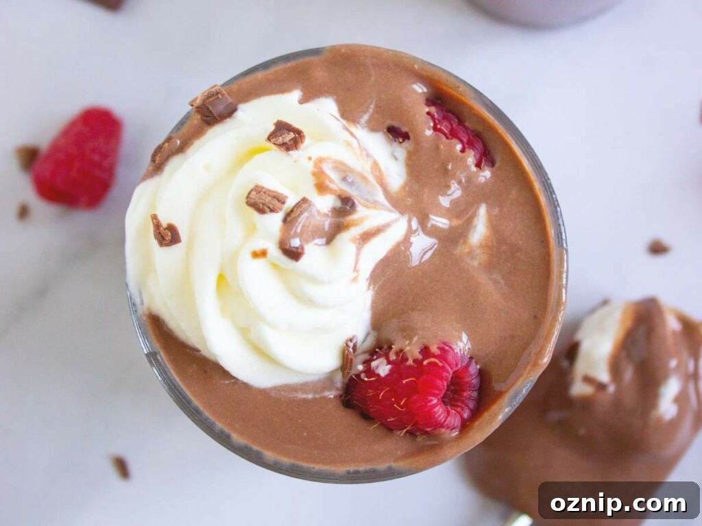 Low Carb Sugar Free Protein Pudding Recipe 3 A glass of protein pudding with whipped cream, chocolate shavings, and raspberries on a white surface.