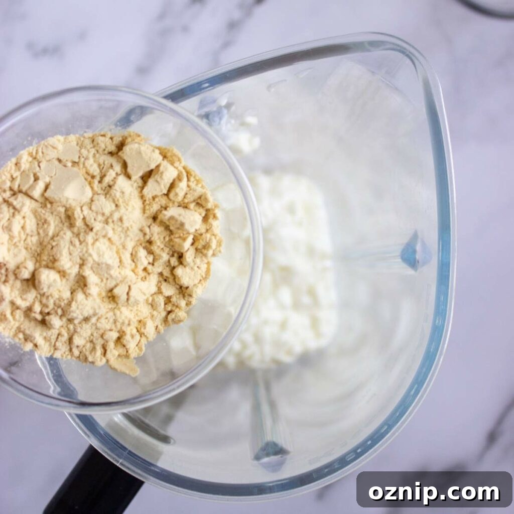 Low Carb Sugar Free Protein Pudding Recipe 7 Protein powder is being poured into a blender containing cottage cheese on a marble countertop.