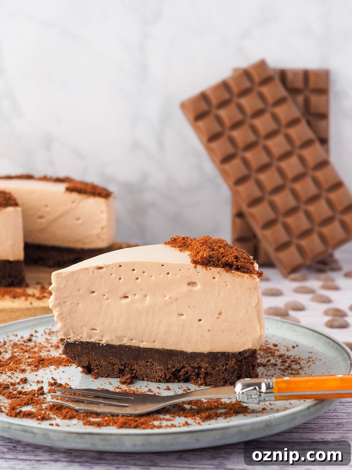 Slice of cheesecake on a plate with a fork and rest of cheesecake and block of chocolate in the background.
