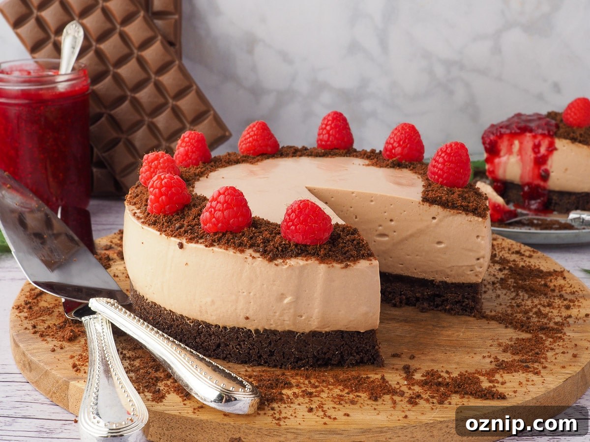 Cheesecake on a serving board, garnished with raspberries, with serving wear and slice of cheesecake, raspberry compote and blocks of chocolate in the background.