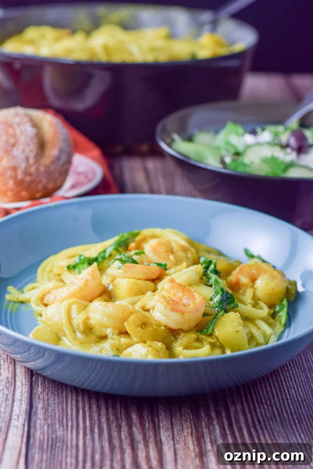 Shrimp with curry sauce and noodles in front of a pan, bread and bowl of salad